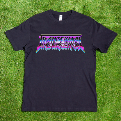 The Transexual Insurgency local hand-printed T-shirt features bold, colorful angular lettering on black fabric, shown laid flat on bright green grass.