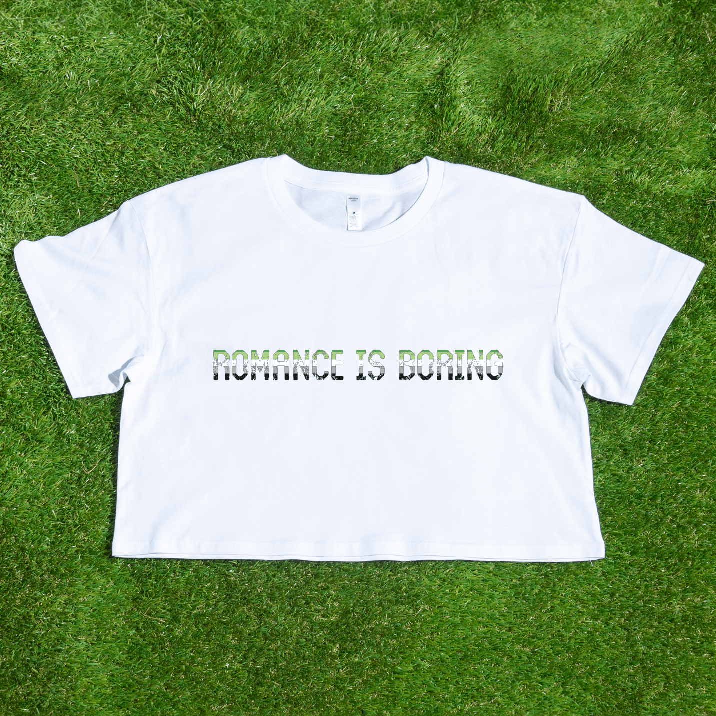 A black crop tee with the text "ROMANCE IS BORING" in green and white lettering is displayed on lush green grass. Celebrate love your way with this unique piece, "Romance Is Boring aromatic aro pride," from Gay Skate, a queer and trans-owned business.