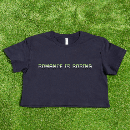 A black crop tee from Gay Skate lies on green grass, featuring the bold white text "ROMANCE IS BORING," celebrating aromatic aro pride from a queer and trans-owned brand.