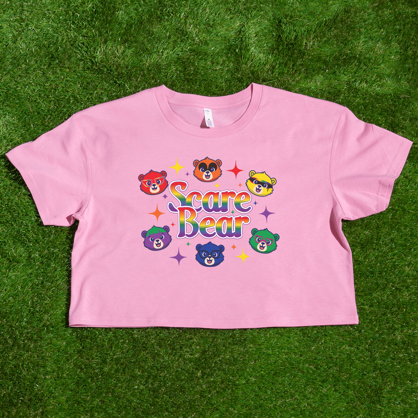 A pink crop top on grass shows colorful cartoon bears and "Scare Bear" in bold letters, surrounded by stars—perfect for adding the SCARE BEAR fundraiser tee, a local hand-printed t-shirt, to your collection.