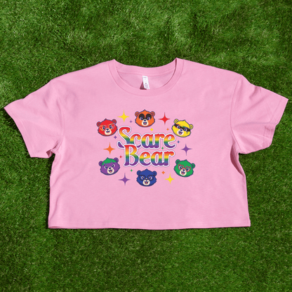 A pink crop top on grass shows colorful cartoon bears and "Scare Bear" in bold letters, surrounded by stars—perfect for adding the SCARE BEAR fundraiser tee, a local hand-printed t-shirt, to your collection.