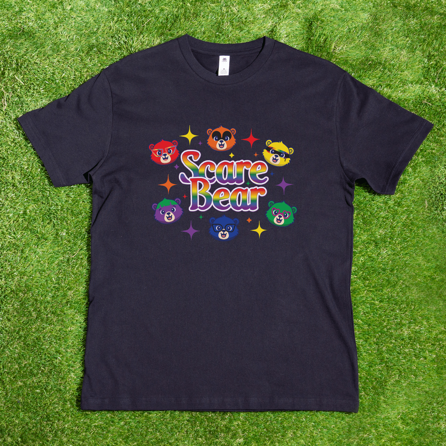 A black SCARE BEAR fundraiser tee—a locally hand-printed t-shirt—lies on grass with colorful cartoon bear faces and rainbow "Scare Bear" text; perfect for roller derby, GAY SKATE, or showing your pride.