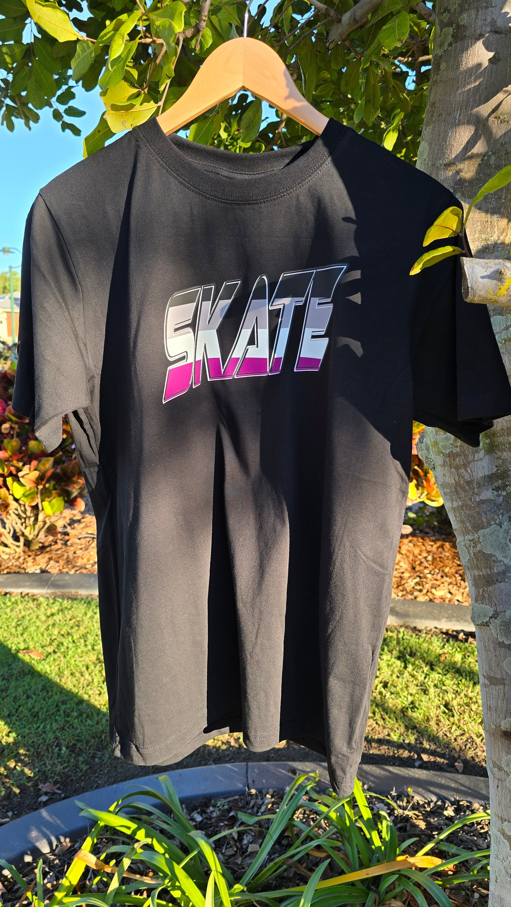 A black "SKATE Asexual flag" t-shirt by Gay Skate, custom-designed and hanging on a tree branch, features the word "SKATE" in bold, gradient white-to-pink text with a dynamic, shadowed design. Sunlight highlights parts of the shirt while colorful foliage and green grass create a vibrant background.