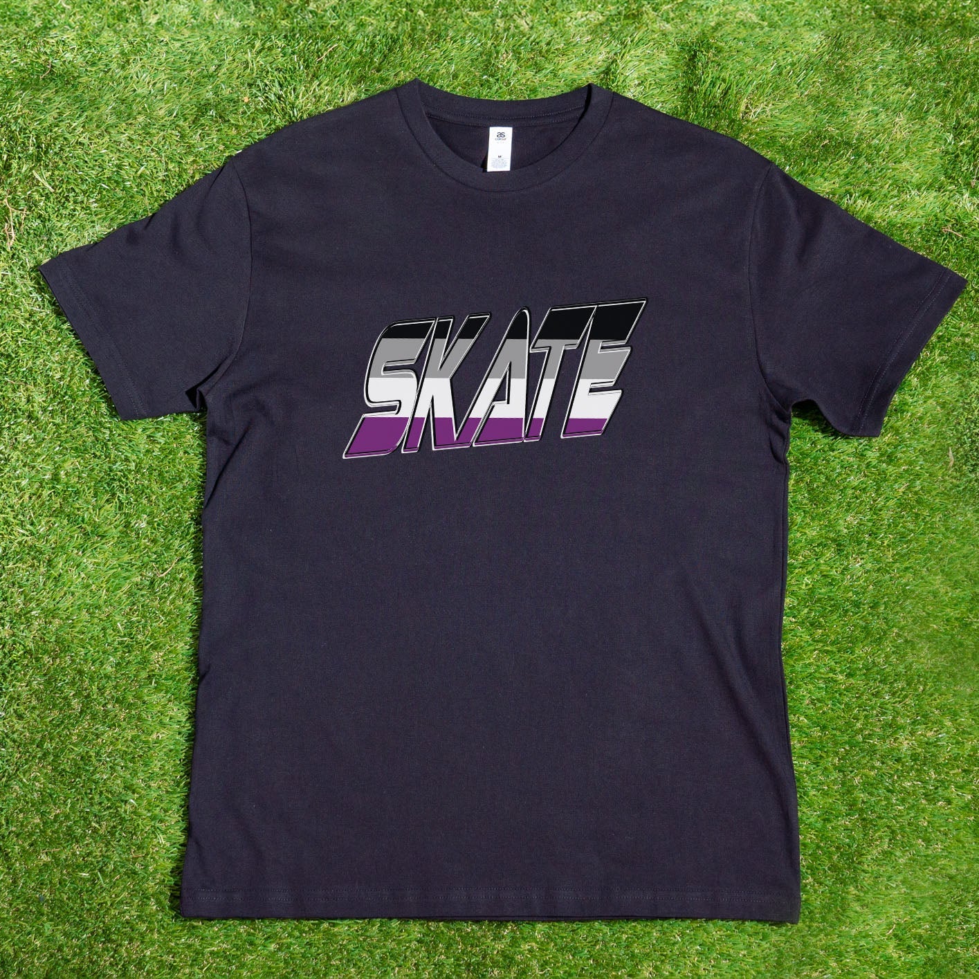 A black "SKATE Asexual flag" t-shirt by Gay Skate, custom-designed and hanging on a tree branch, features the word "SKATE" in bold, gradient white-to-pink text with a dynamic, shadowed design. Sunlight highlights parts of the shirt while colorful foliage and green grass create a vibrant background.