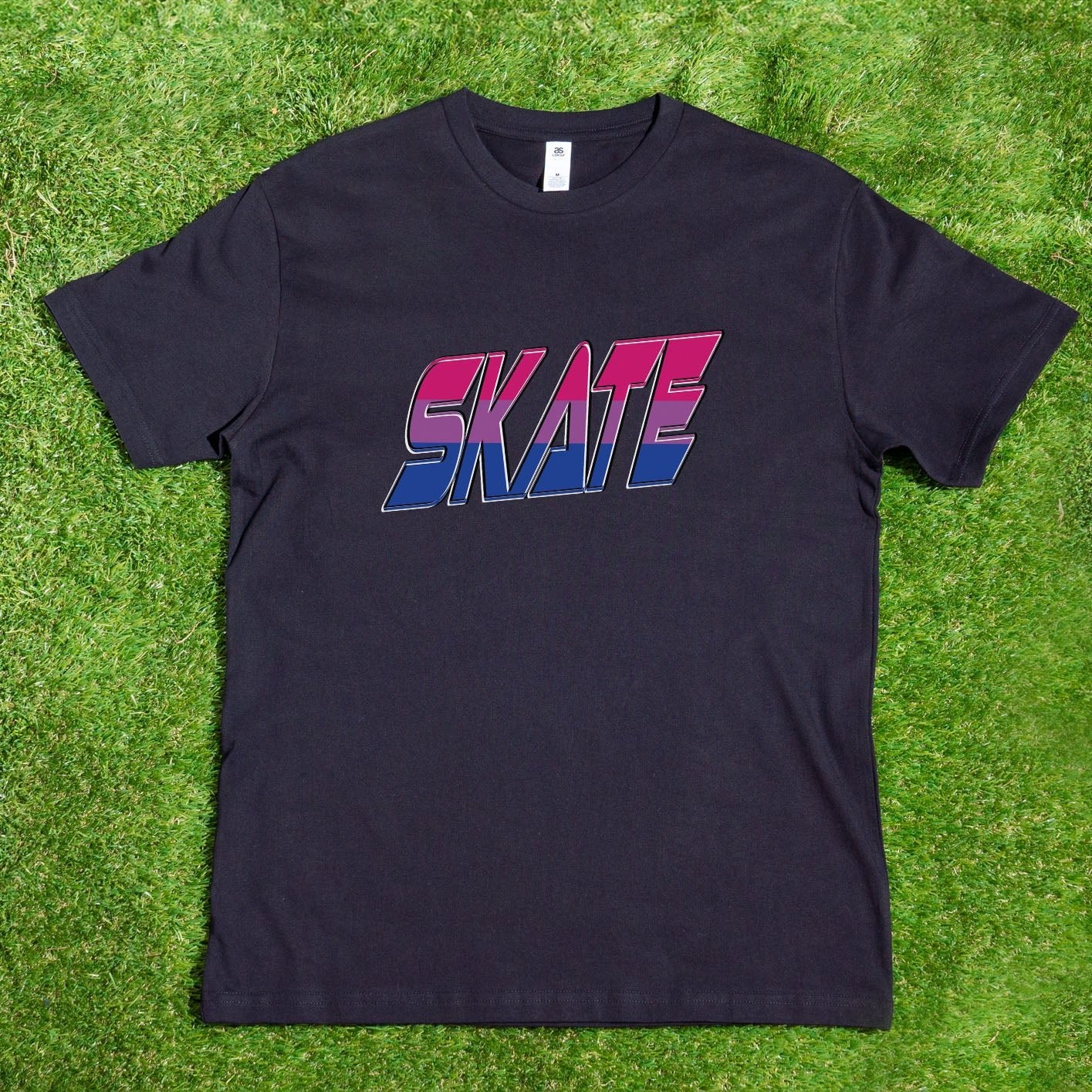 A SKATE Bisexual - Local hand printed t-shirt from My Store in black, adorned with the word "SKATE" in bold, colorful letters, hangs on a wooden hanger from a tree branch. The background features a lush garden with green grass, vibrant plants, winding pathways, and a glimpse of a house. Sunlight brightly illuminates the scene, reflecting My Store's support for diversity and custom orders.