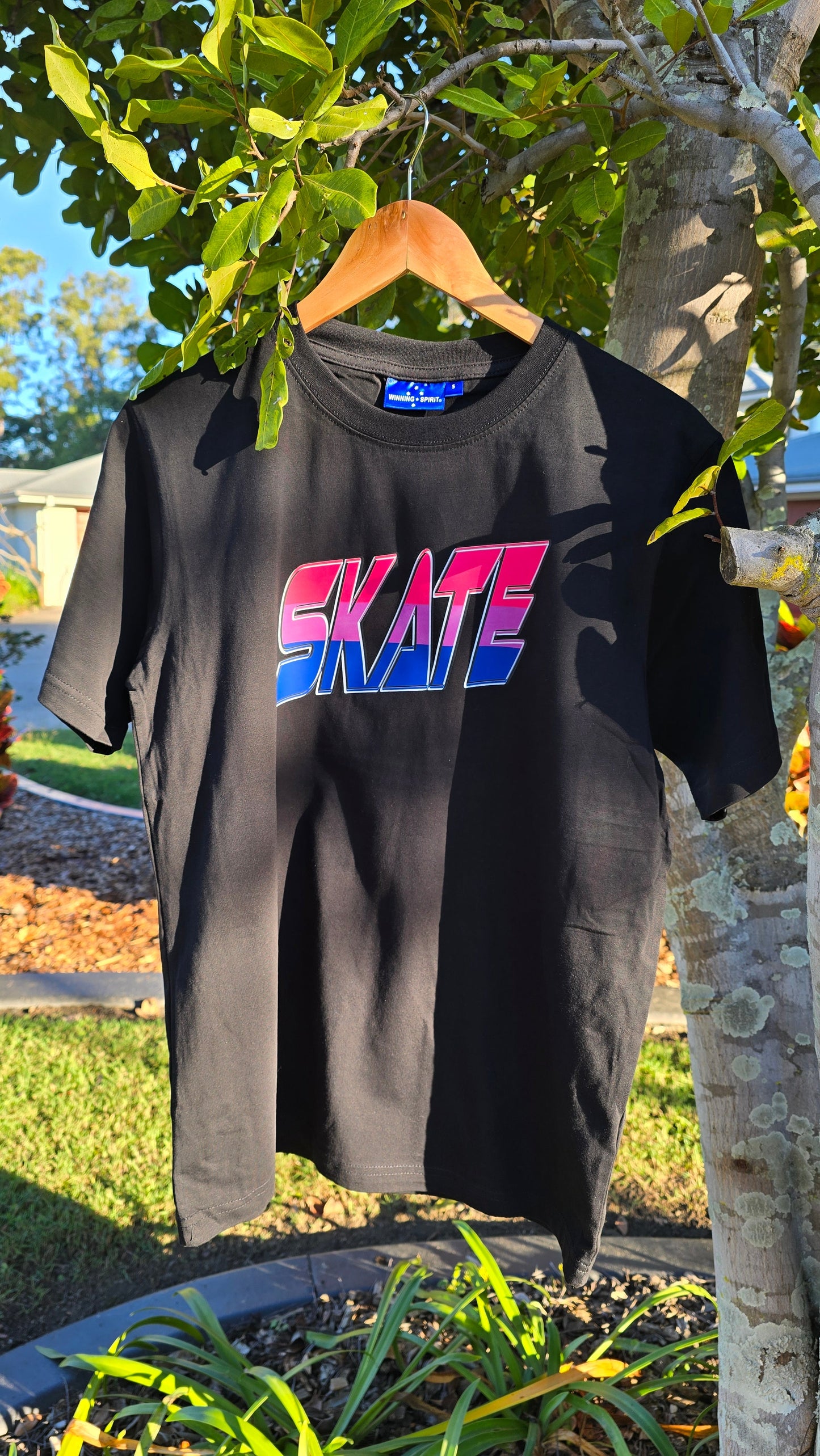 A SKATE Bisexual - Local hand printed t-shirt from My Store in black, adorned with the word "SKATE" in bold, colorful letters, hangs on a wooden hanger from a tree branch. The background features a lush garden with green grass, vibrant plants, winding pathways, and a glimpse of a house. Sunlight brightly illuminates the scene, reflecting My Store's support for diversity and custom orders.