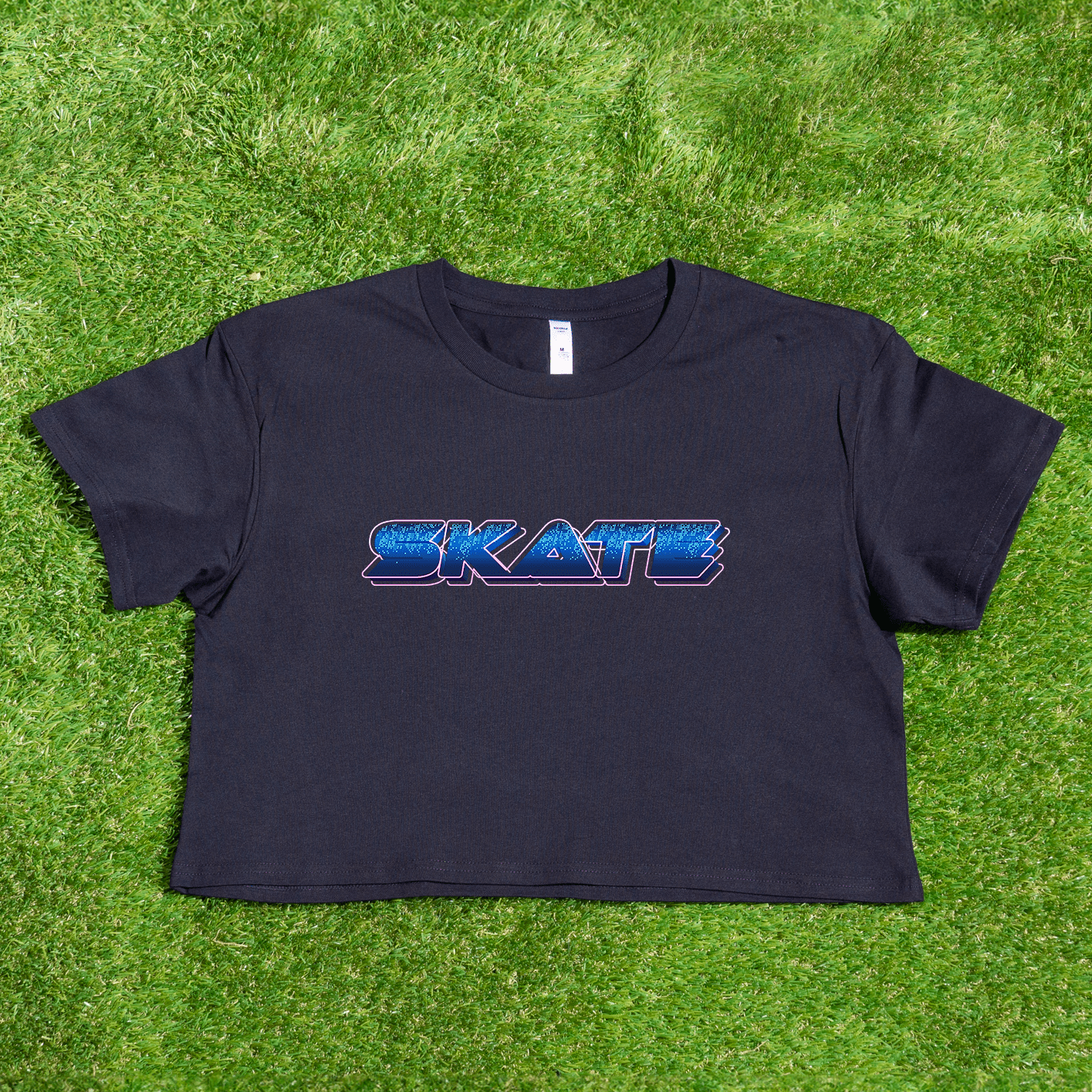 A black crop tee from Gay Skate, featuring the "SKATE" blue cyber logo, is laid flat on green grass, showcasing the vibrant essence of queer skate streetwear.