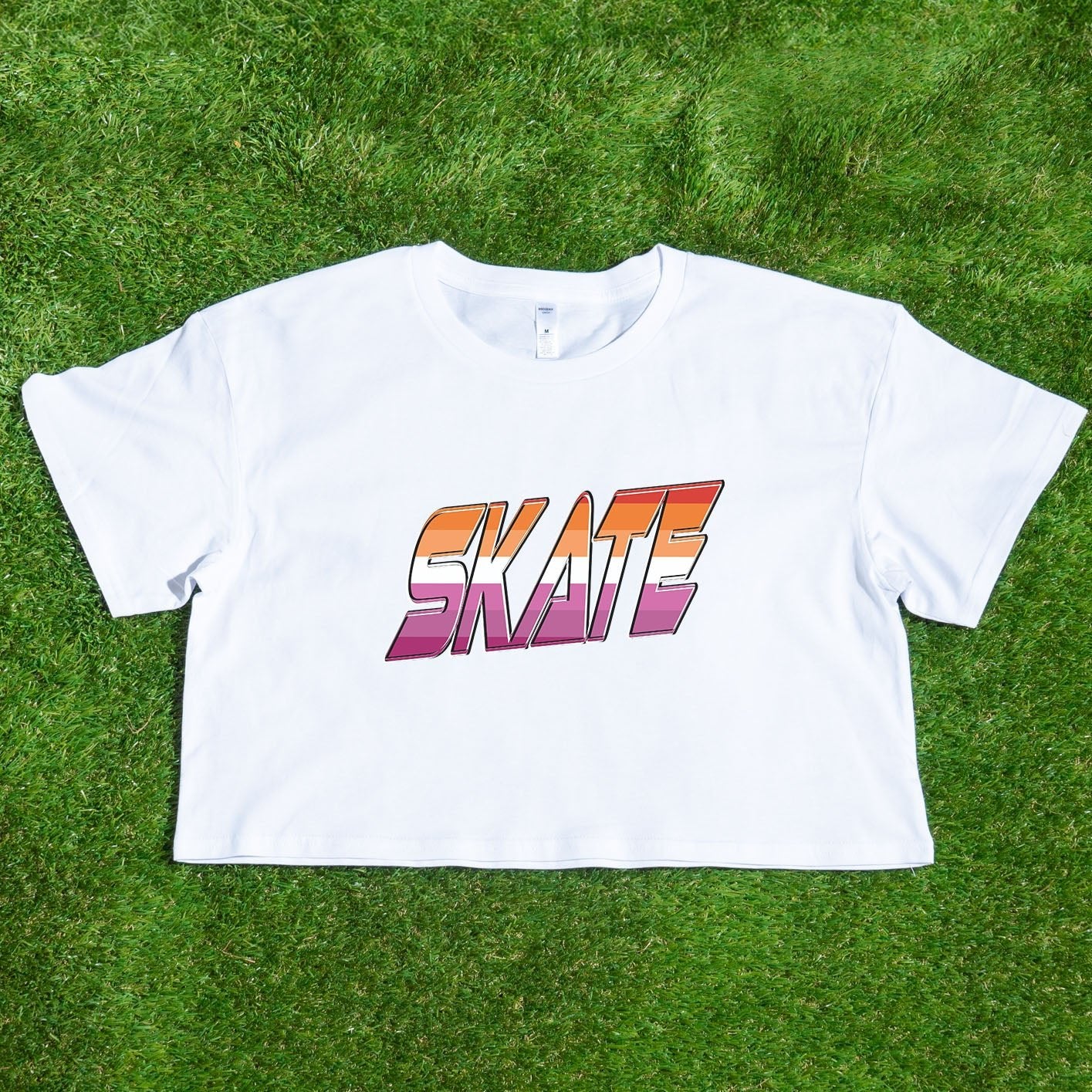 Gay Skate's SKATE lesbian pride flag - locally printed crop tee is a white cropped shirt featuring the word "SKATE" in bold, gradient letters transitioning from orange to pink. The retro, 3D-style lettering adds a distinct flair to this queer skate streetwear piece, which also includes short sleeves and a round neckline.