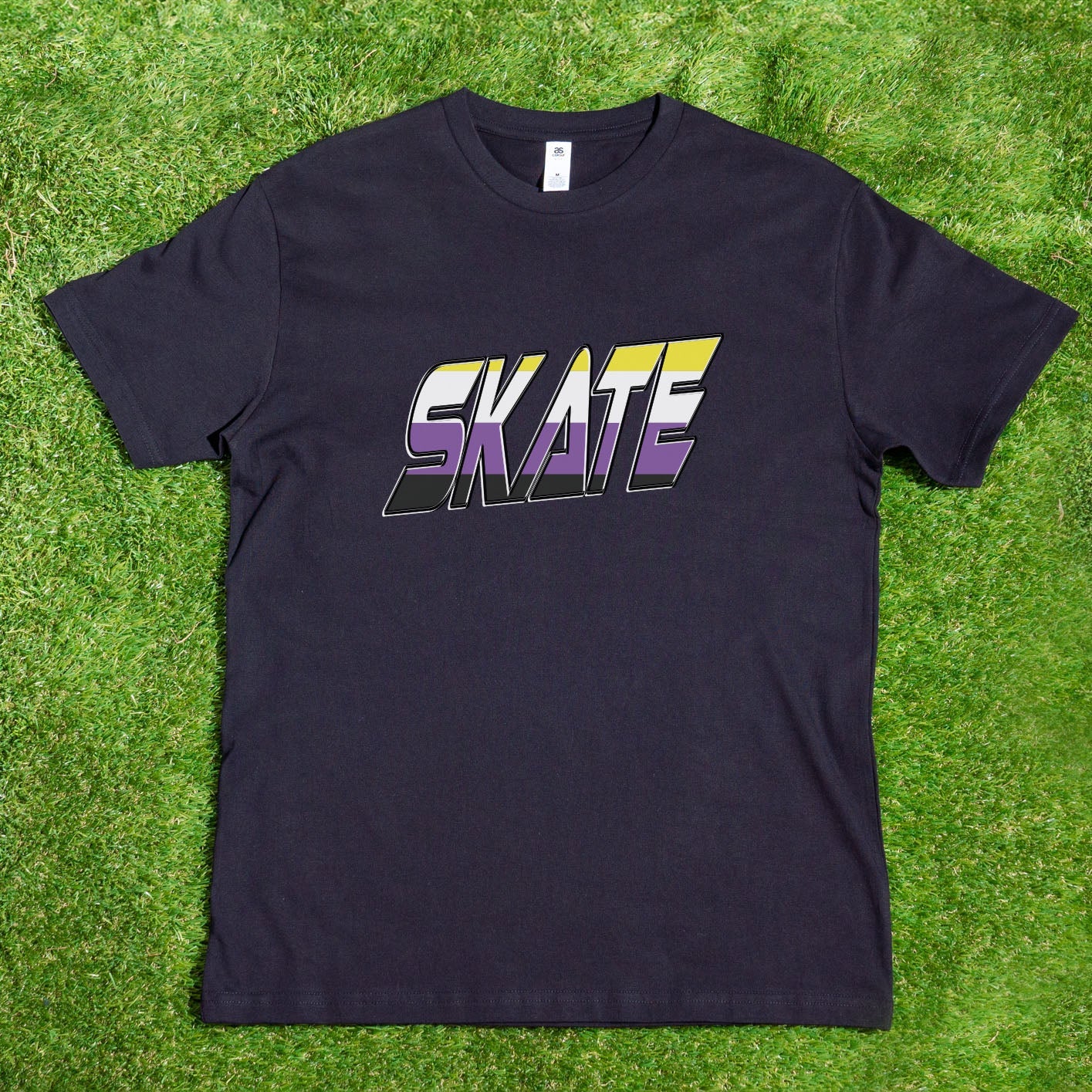 The "SKATE Non-Binary Flag - Local hand printed t-shirt" from My Store, displayed outdoors hanging on a tree branch, showcases the word "SKATE" in large, bold letters adorned with a stunning gradient of yellow, purple, and blue. This vivid design celebrates diversity and stands out beautifully against the lush greenery on a sunny day.