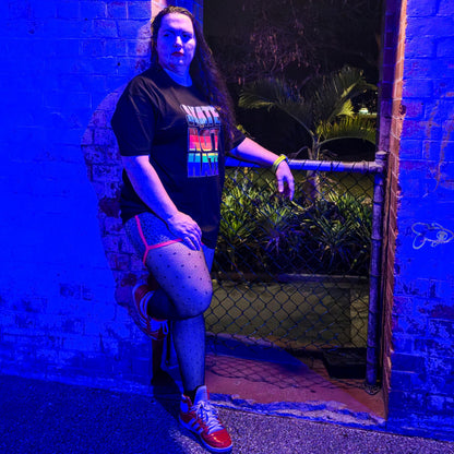 A person wearing a black “Skate Not Hate - Local hand printed t-shirt” and red shoes poses by a brick doorway at night, illuminated by blue light, with greenery visible behind a chain-link gate.