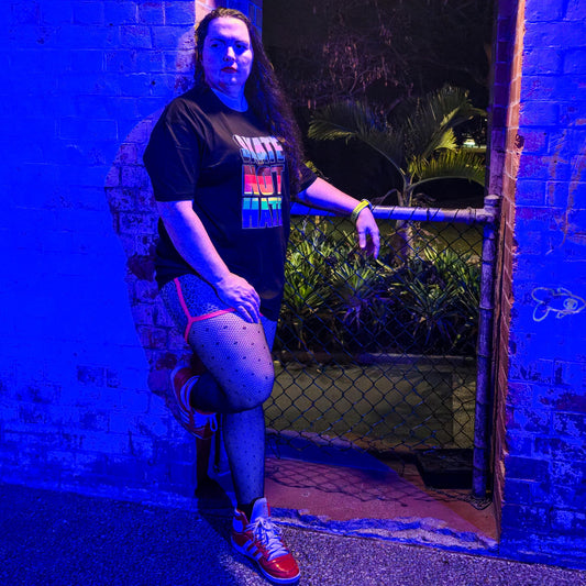 A person wearing a black “Skate Not Hate - Local hand printed t-shirt” and red shoes poses by a brick doorway at night, illuminated by blue light, with greenery visible behind a chain-link gate.