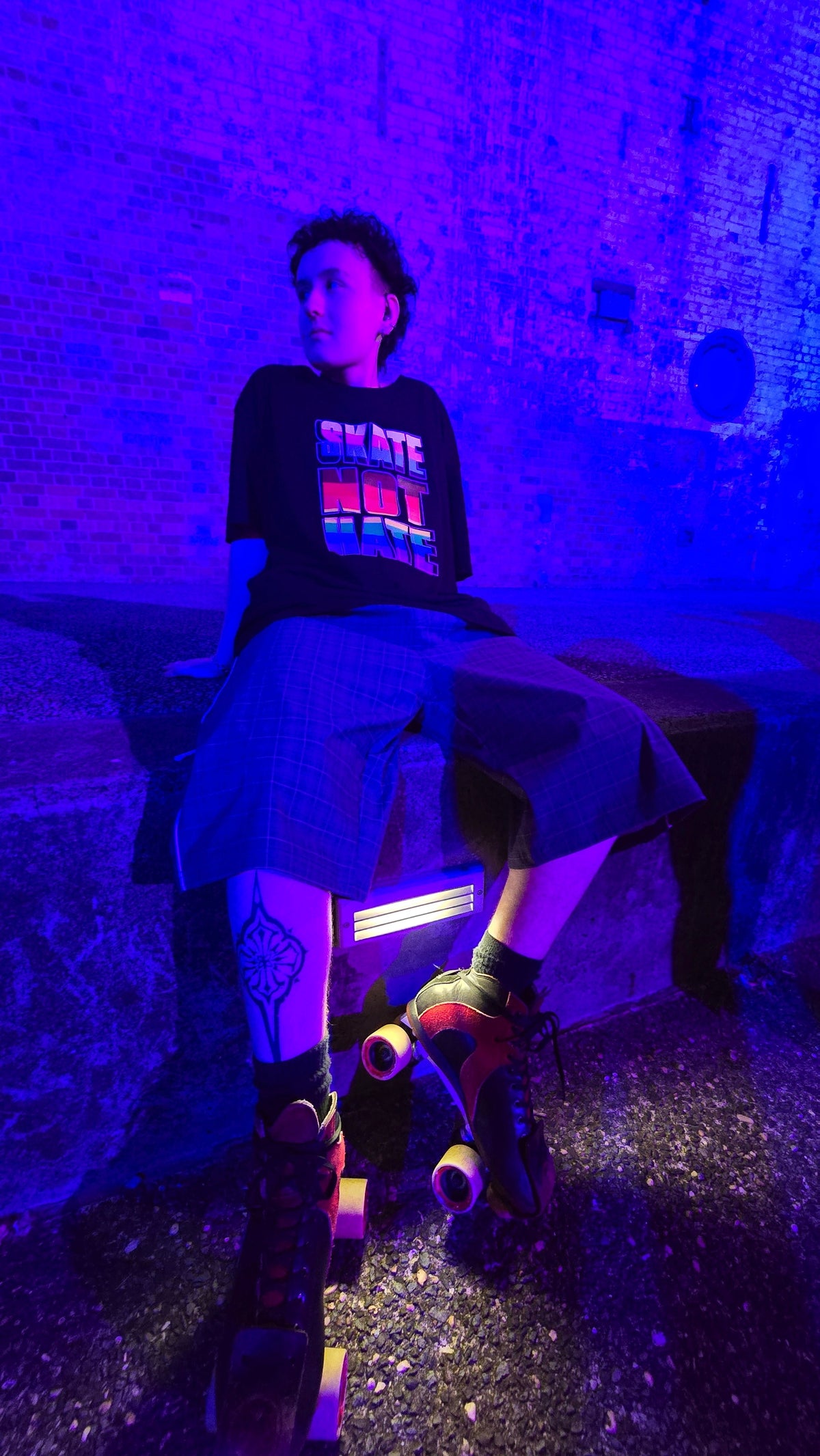 Nico wearing a black graphic SKATE NOT HATE t-shirt, long gray pants, and roller skates sits on a bench in a dimly lit urban setting under strong blue and purple lighting.