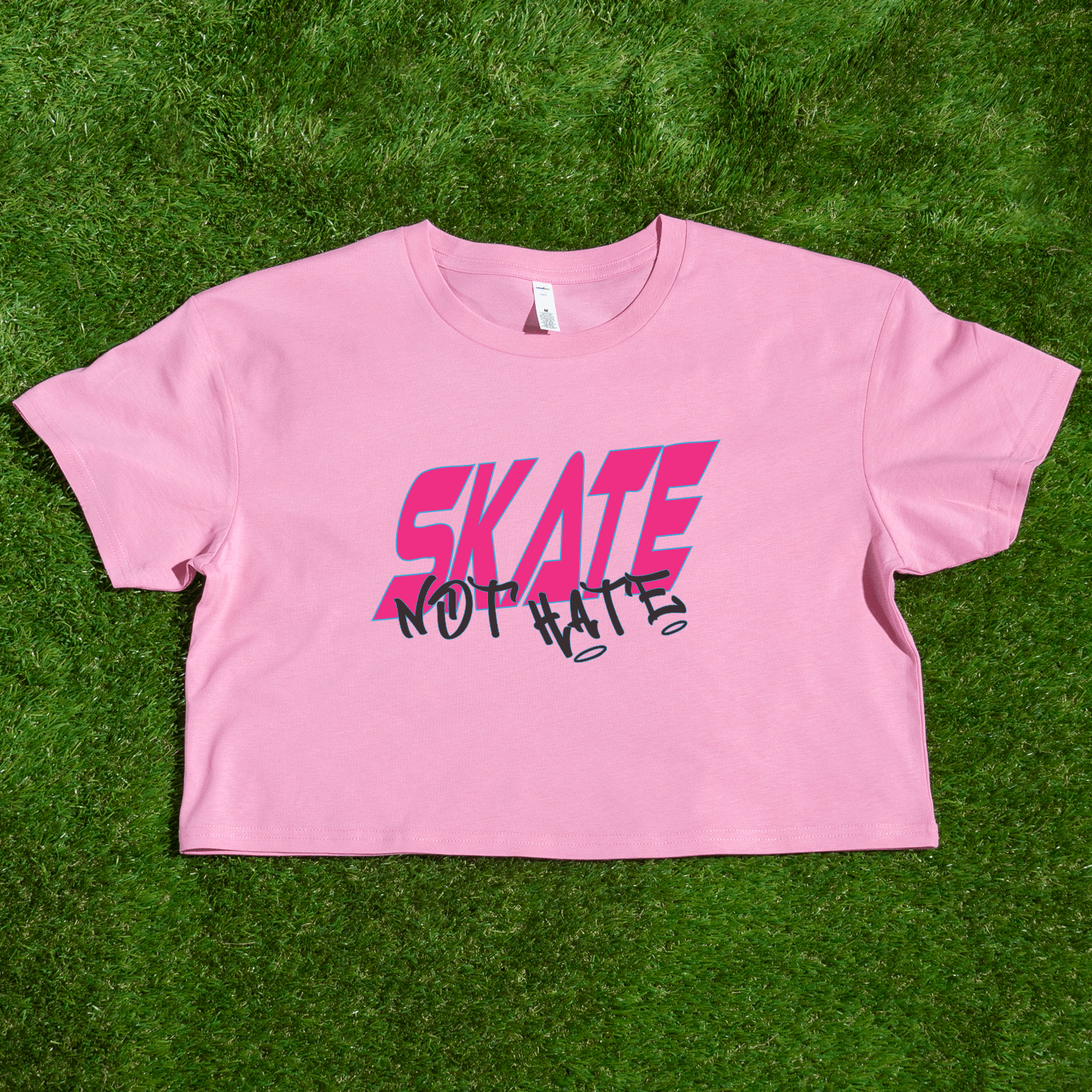 The SKATE NOT HATE original logo crop tee by Gay Skate is a white, short-sleeved shirt featuring the word "SKATE" in bold, pink, slanted capital letters across the chest. The message is accentuated with the word "NOT" in black, distorted text overlaying "SKATE," promoting the message: skate not hate.