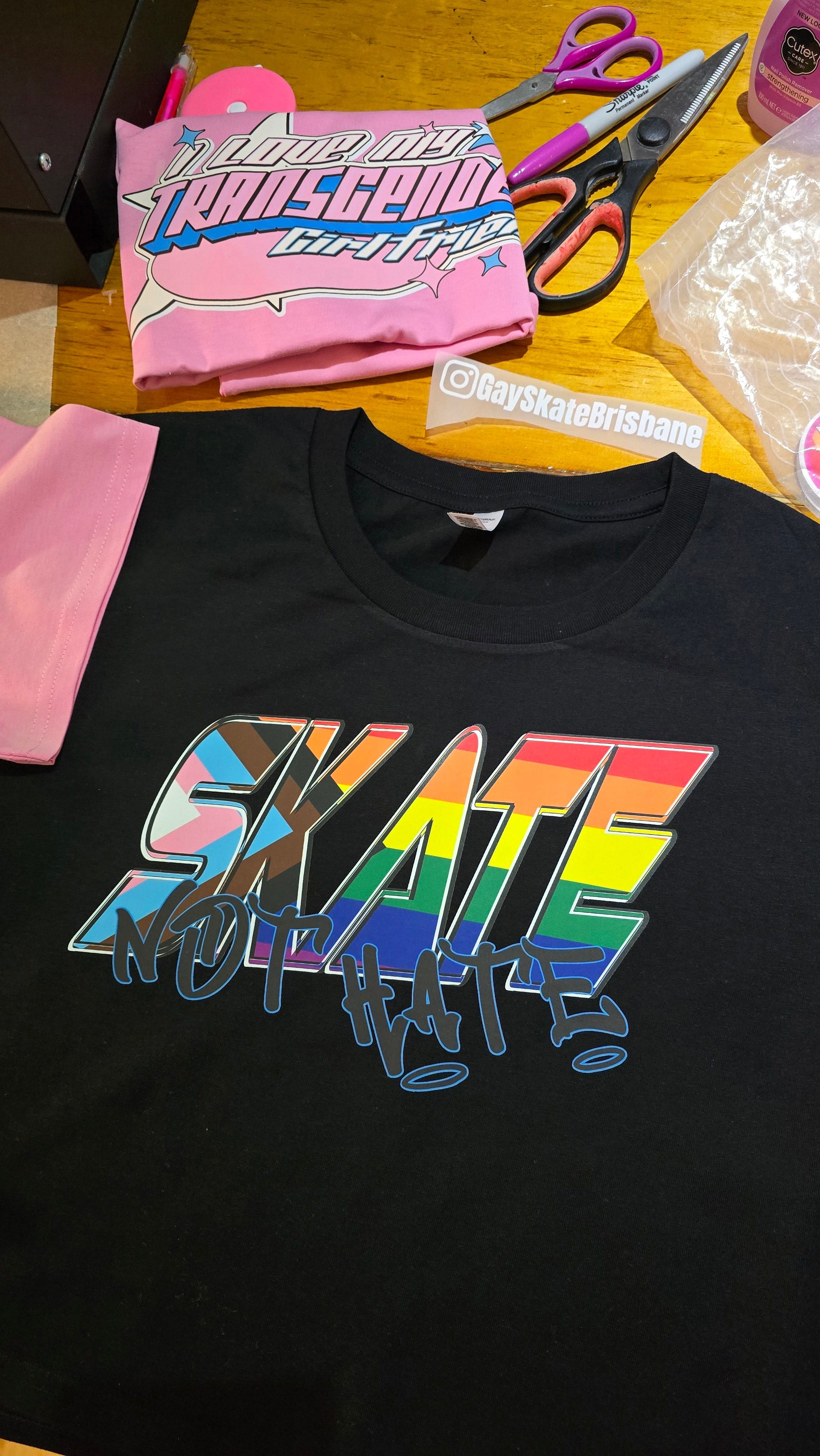 The SKATE NOT HATE original logo crop tee by Gay Skate is a white, short-sleeved shirt featuring the word "SKATE" in bold, pink, slanted capital letters across the chest. The message is accentuated with the word "NOT" in black, distorted text overlaying "SKATE," promoting the message: skate not hate.