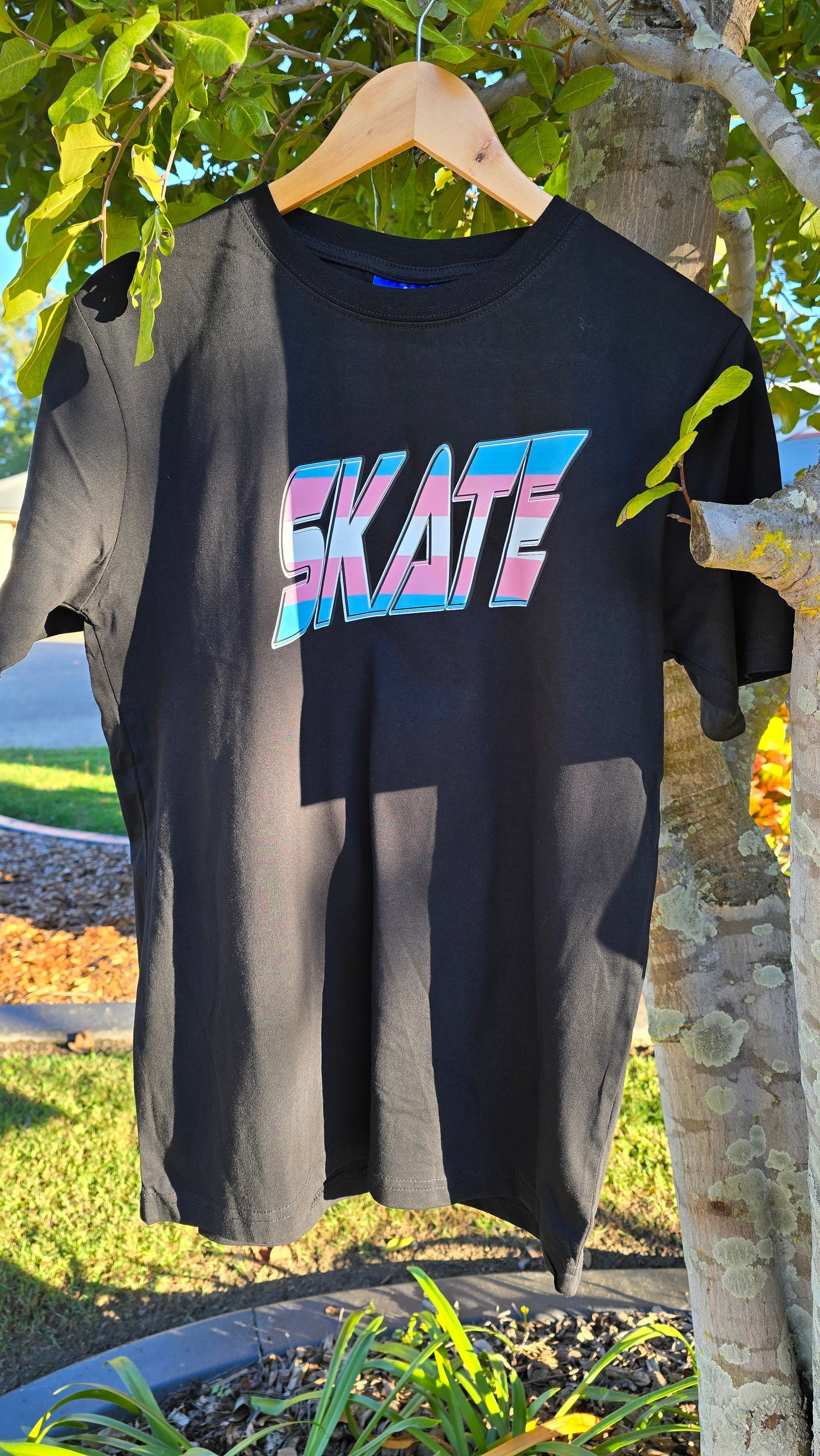 A black My Store T-shirt, featuring the word "SKATE" printed in large, bold gradient letters in pink, blue, and white shades from the Trans Flag design, hangs on a wooden hanger attached to a tree branch. The background displays lush greenery and a winding pathway in a sunny outdoor setting, subtly celebrating support for diversity.