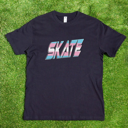 A black My Store T-shirt, featuring the word "SKATE" printed in large, bold gradient letters in pink, blue, and white shades from the Trans Flag design, hangs on a wooden hanger attached to a tree branch. The background displays lush greenery and a winding pathway in a sunny outdoor setting, subtly celebrating support for diversity.