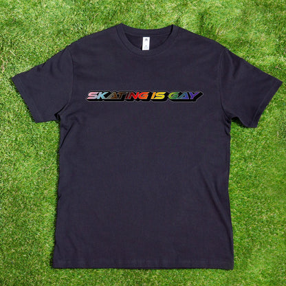 A black "Skating Is Gay - Local Hand-Printed T-Shirt" from My Store, crafted from 100% carded cotton, hangs on a wooden hanger attached to a tree branch. The shirt proudly displays the phrase "SKATING IS GAY" in vibrant rainbow letters across the chest, celebrating gay skating culture. The outdoor setting is adorned with lush greenery and sunlight casting playful shadows.