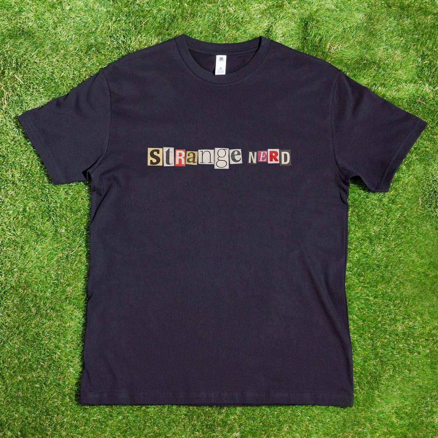 A black Gay Skate T-shirt lies on the grass, proudly displaying "Strange Nerd Transgender Anagram" in multicolored, ransom-note style letters across the front.