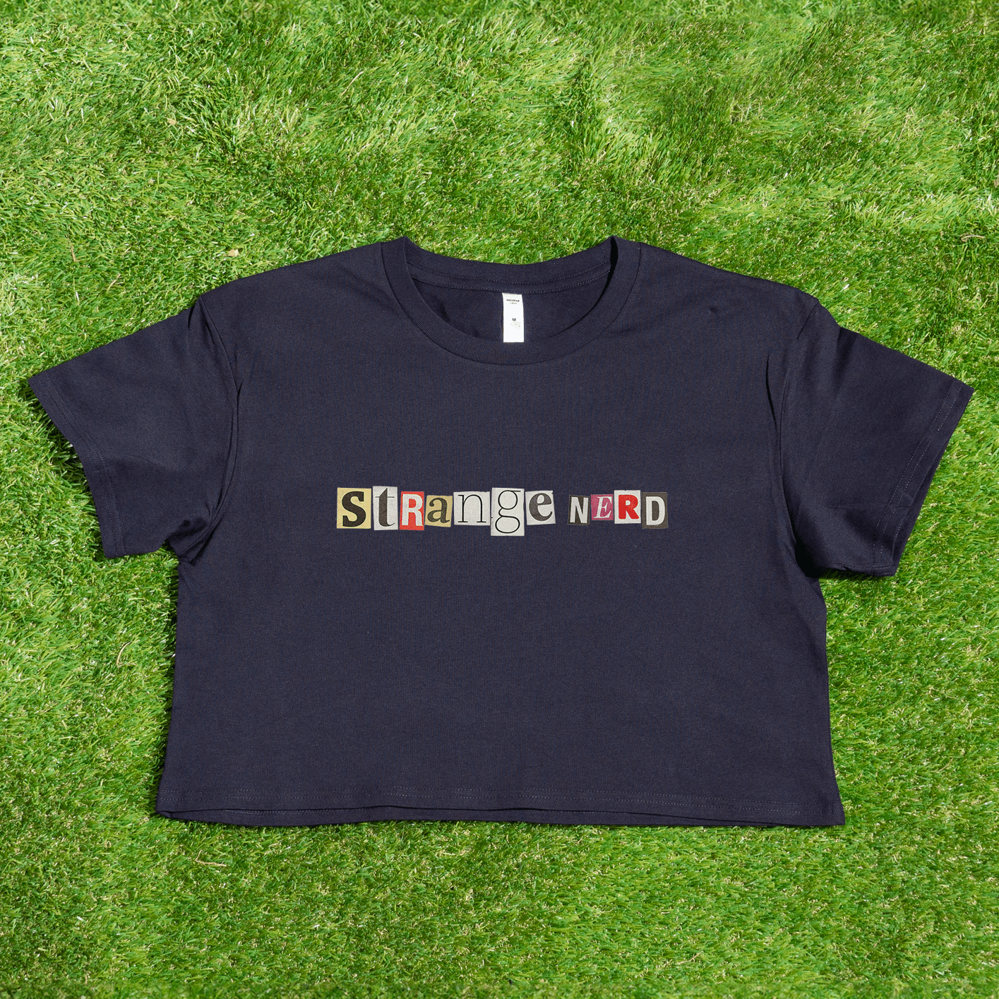 A black cropped tee featuring "STRANGE NERD" in vibrant, ransom-style letters rests on lush green grass, capturing the edgy essence of queer skate streetwear. Celebrate self-expression with this distinctive piece from a trans-owned small business, the Strange Nerd transgender anagram crop tee by Gay Skate.