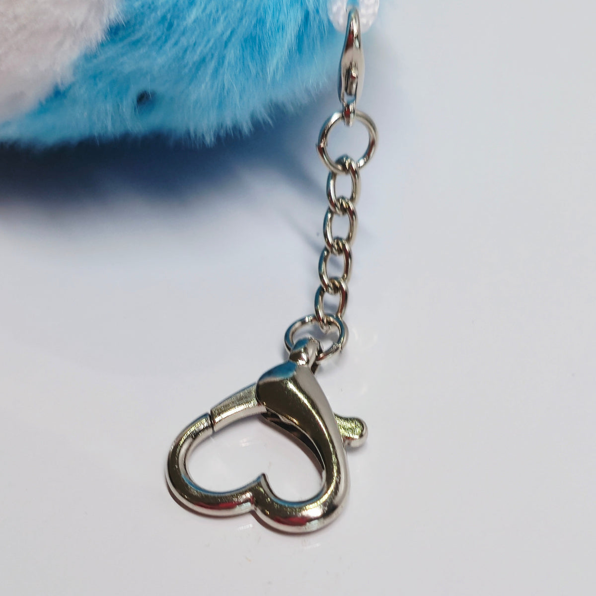 15 cm blue, pink and white trans pride plush shark keychain lying beside its silver-tone heart-shaped carabiner and keyring.