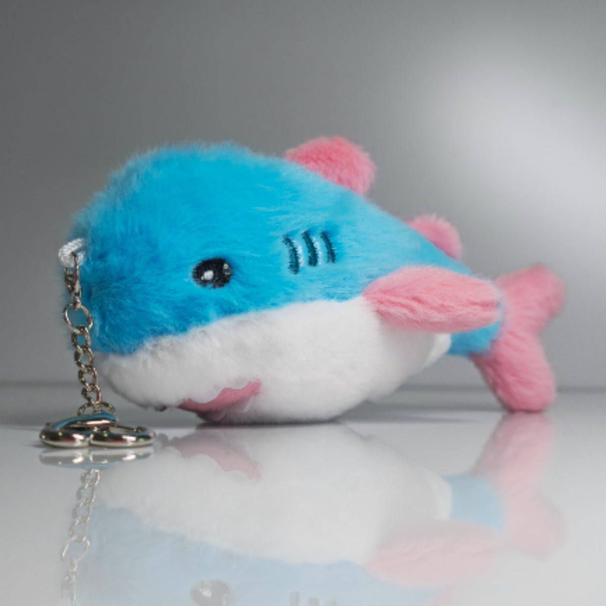 15 cm blue, pink and white trans pride plush shark keychain lying beside its silver-tone heart-shaped carabiner and keyring.