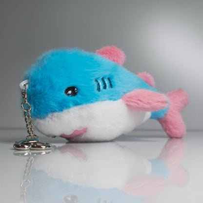 15 cm blue, pink and white trans pride plush shark keychain lying beside its silver-tone heart-shaped carabiner and keyring.