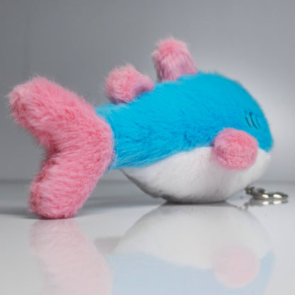 15 cm blue, pink and white trans pride plush shark keychain lying beside its silver-tone heart-shaped carabiner and keyring.