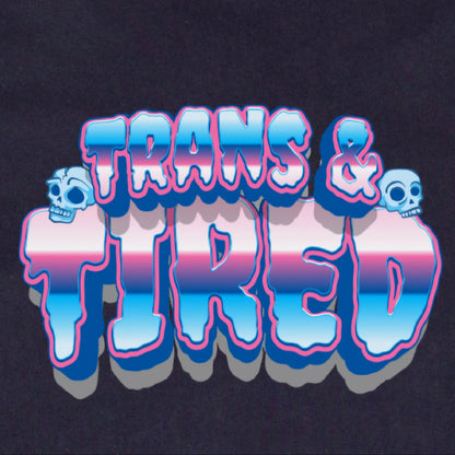 The Trans & Tired - Local hand printed t-shirt features "TRANS & TIRED" in bold, 3D letters with blue, pink, and white stripes plus two cartoon skulls on a black background inspired by the transgender pride flag.