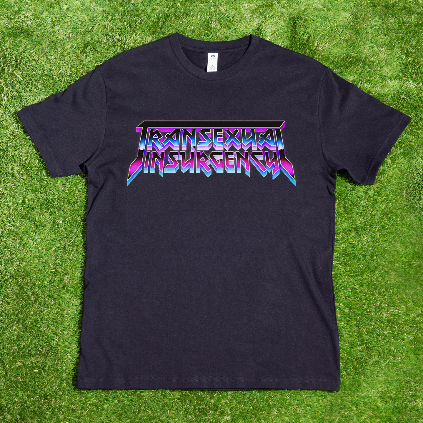 The Transexual Insurgency local hand-printed T-shirt features bold, colorful angular lettering on black fabric, shown laid flat on bright green grass.