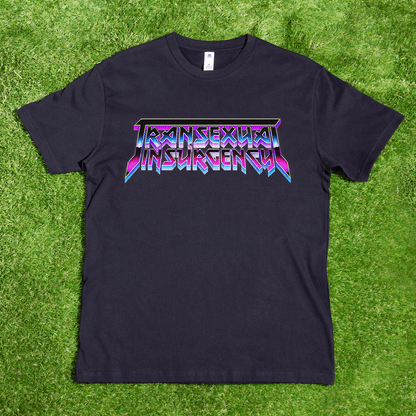 The Transexual Insurgency local hand-printed T-shirt features bold, colorful angular lettering on black fabric, shown laid flat on bright green grass.