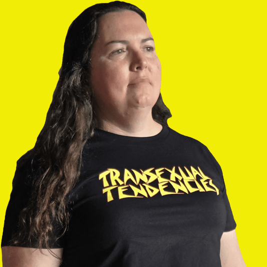 A person with long brown hair wears the TRANSEXUAL TENDENCIES local hand printed satire t-shirt, featuring bold yellow lettering on a black crop top, standing against a vibrant yellow background—a striking queer pride statement.