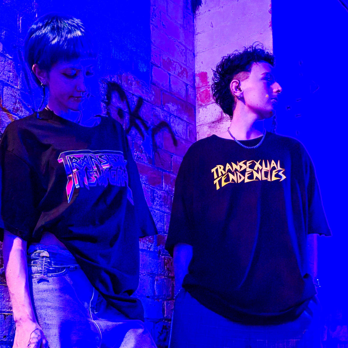 Two people stand against a graffiti-covered brick wall under blue-purple lighting, both wearing oversized TS TENDENCIES local hand printed satire t-shirts with bright logos that read “TRANSEXUAL TENDENCIES.”.