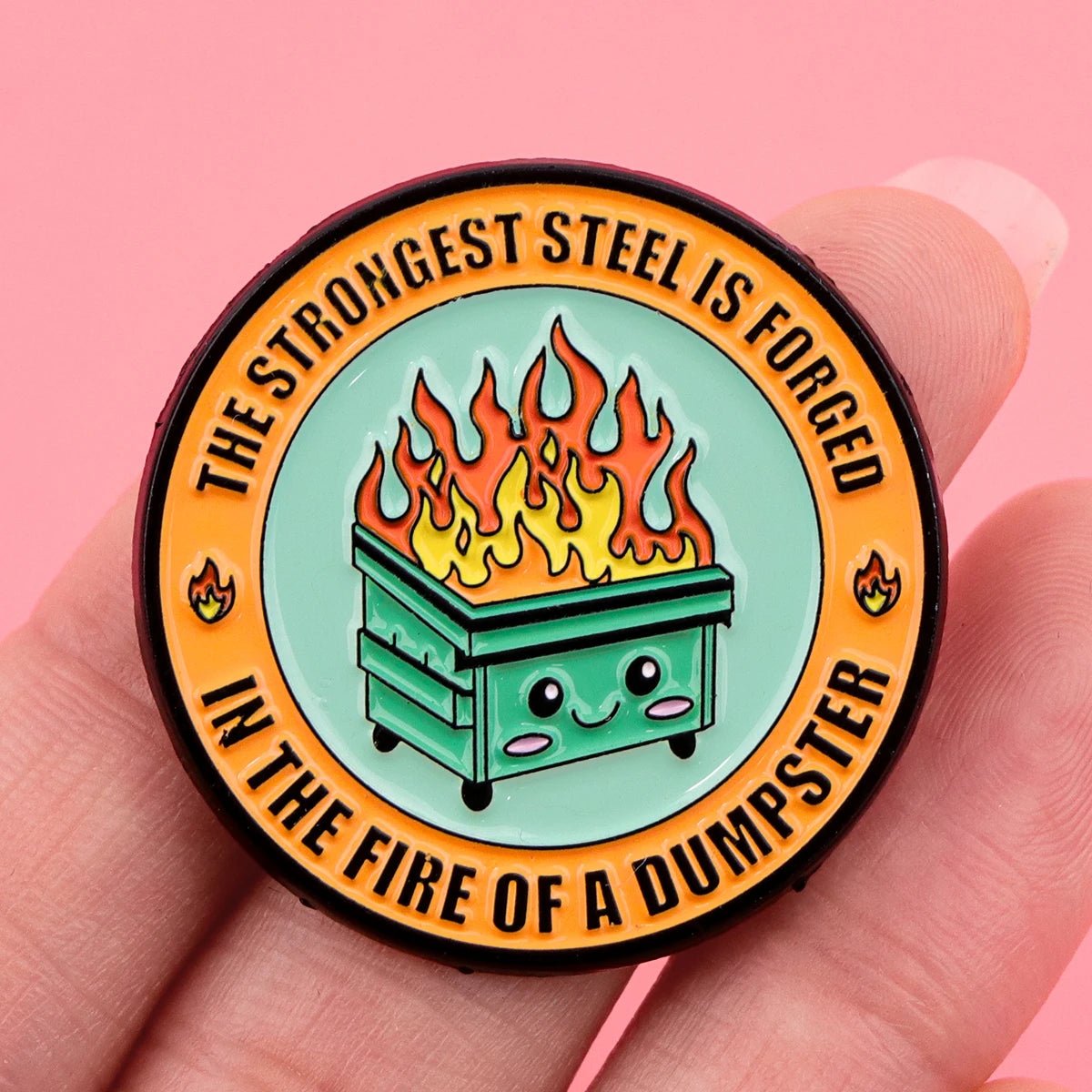 The "Very Demure Very Mindful Very Cutesy" enamel badge boasts a cute, smiling dumpster on fire against a teal backdrop with an orange border that reads, "THE STRONGEST STEEL IS FORGED IN THE FIRE OF A DUMPSTER." It stands out beautifully when held by hand against a pink background with a bow accent.