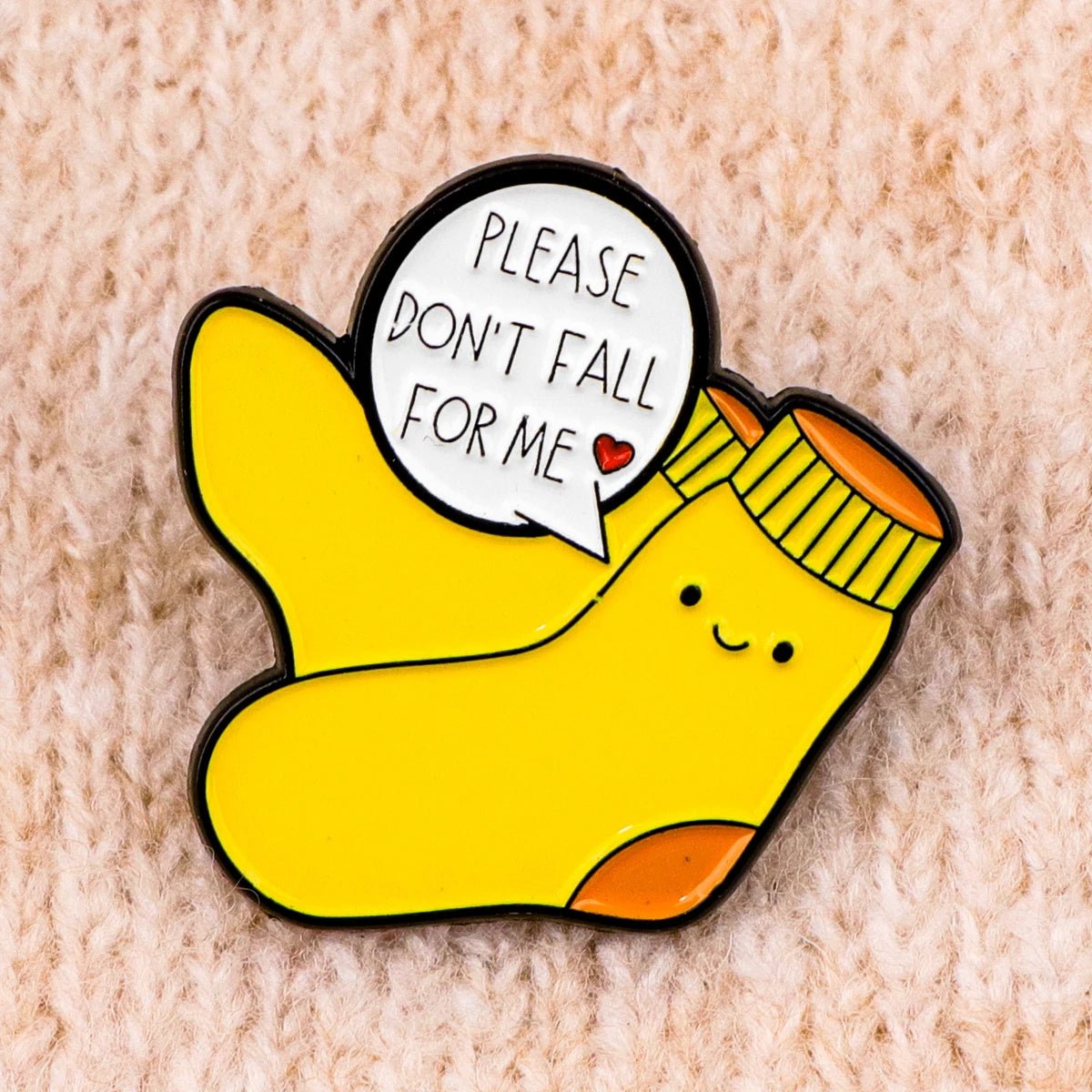 The "Very Demure Very Mindful Very Cutesy" enamel badge features a yellow sock design with orange toes and heels, adorned with a pink bow. A speech bubble says, "Please don't fall for me," alongside a red heart, all set against a textured beige fabric background, making it truly stand out.