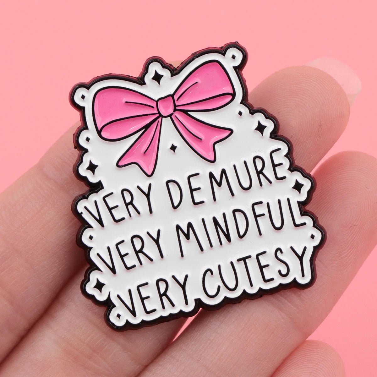 The "Very Demure Very Mindful Very Cutesy" enamel badge showcases a pink bow with the text on a pink background, adding playful elegance to any accessory collection.