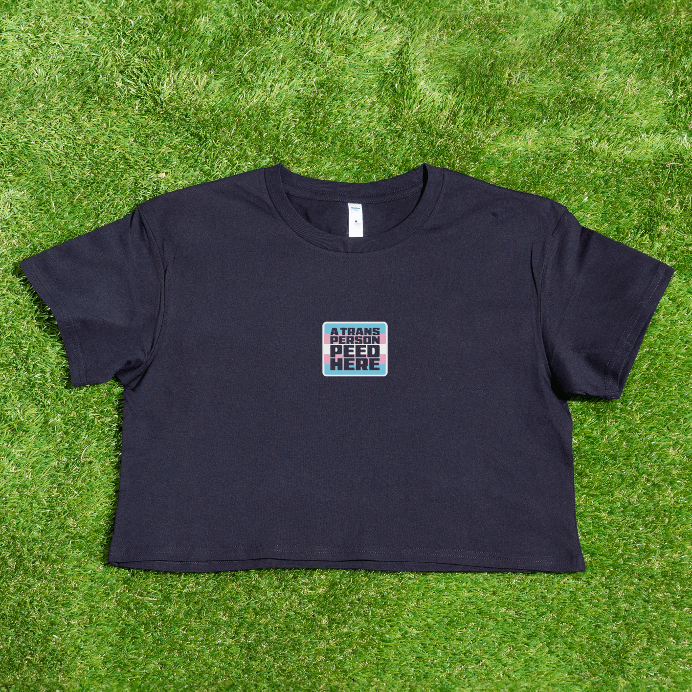 Black crop t-shirt with a small square logo: A Trans Person Peed Here featuring trans flag
