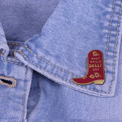 A red cowboy boot-shaped enamel pin with gold lettering that reads 'WHAT WOULD DOLLY DOT' and small gold floral details, pinned to a light blue denim jacket.